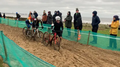 Three riders have been forced to get out of their saddles and walk through a section of deep sand. Spectators are cheering them on at the side of the course.