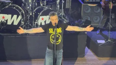 Robbie Williams in a black Slade T-shirt is on stage with his arms outstretched at his sides