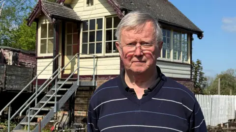Alan Jones, a man wearing glasses and a dark blue jumper with thin white stripes and thin light blue stripes. He is standing in front of an old-fashioned signal box.