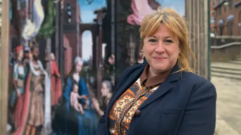 A woman with shoulder length hair, wearing a blue suit jacket and patterned top looks to the camera. She is stood to the right of a large painting, which is slightly blurred