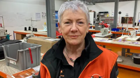Carole Champion is standing in a factory. She is wearing a black t-shirt and a black and orange jacket. In the background there are sewing machines on workbenches. 