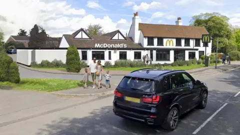 A generated image of a black car driving past what the McDonald's building could look like while families walk on the pavement