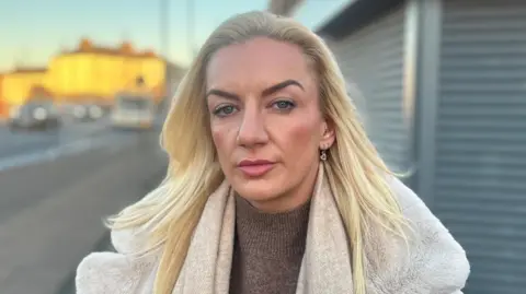 BBC Amy is in her mid-thirties and has long, dyed blonde hair and is wearing a cream coat. She is standing next to a Shoppes roller blinds which are out of focus behind her.