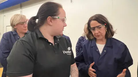 Emma Baugh/BBC Liz Kendall is wearing dark blue overalls and safety glasses, she is speaking with a woman wearing a black polo shirt and safety glasses, behind her are several people also wearing overalls, they are in a room with white walls.