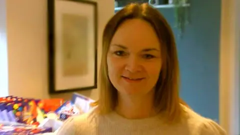 Laura Evans wearing a white jumper and smiling at the camera. She has shoulder-length dark blonde hair and there are presents behind her 