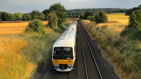 A photo of a train going through a field, there is a train in the middle on the tracks. It is a Chiltern train and you can see it's white on the top and yellow at the front. 