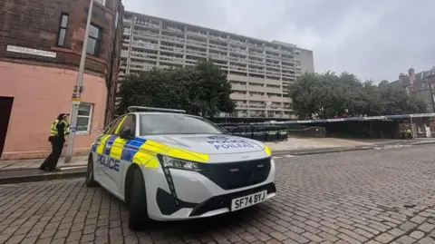 A cobbled street cornered off with police tape as a uniformed officer stands guard next to a police vehicle 
