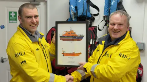 RNLI/ Darren Blight Neil Humphrey presenting Steve Bennetts a framed picture of Plymouth's all-weather lifeboat Sybil Mullen Glover and previous inshore lifeboat Millennium Forester. Both men are wearing yellow RNLI jackets and are shaking hands.