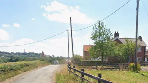 A road with a red brick farm building to the right with barns in the background. 