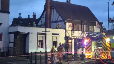 Firefighters at the Black Bear Pub. The road in front of the pub has been cordoned off with red tape and there is a fire engine parked on the road. There are two fire fighters outside of the pub and three on the roof. 