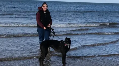 Herout of Lotte's Lotte Stand with his dog, black friend, in the shallow waves of the beach. He has shoes, blue jeans rolled, and wear dark gils on Hooon Moune.