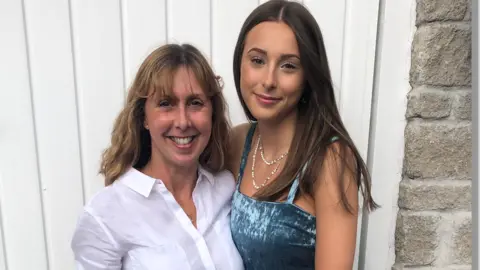 Wearing a white shirt, Debbie Hollinson is seen on the left, smiling, while daughter Darcy Hollinson is on the right, with long brown hair, necklaces and a blue velvet dress. 