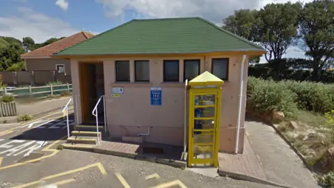A mid shot of the Green Island public toilets. It is a small hut with a green roof, cream coloured walls, and a yellow phone box outside.