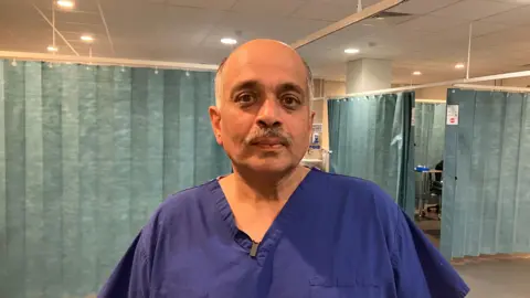 Rakesh Kucheria in blue uniform looking at the camera with a hospital ward behind him.