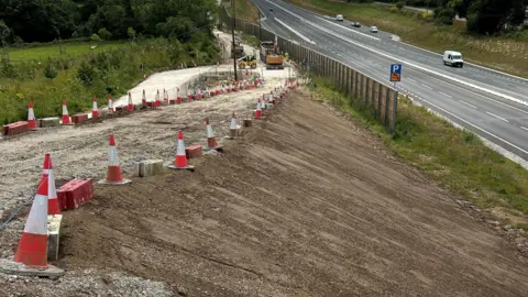 National Highways The side of the A30 which features a large bank of top soil and several traffic cones laid out in a line