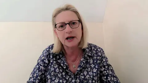 A woman is wearing glasses and a dark blue, patterned top, with a necklace visible at the neckline. She sits in front of a blank beige and white wall.