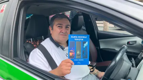 JOANNA TAYLOR/BBC Cenk Sözen holding up his book while sitting in his taxi. The book is a blue paperback with Turkish text and pictures of a taxi, German plane and the aftermath of a bombing. He is wearing a white polo shirt and sitting in the driver's seat. 