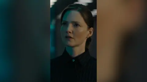 A woman with dark hair pulled back from her face looks at someone off camera, she wears a dark shirt