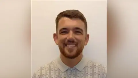 A young man is smiling at the camera. He is wearing a light coloured, polo-style top with a geometric pattern. He has dark hair and a beard.