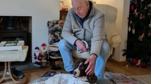 The Thin Blue Paw Foundation John Sexton is stroking Red who is laying on the floor. It is Christmas time in the household with a tree up in the background. 