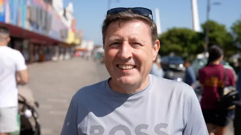 Qays Najm/BBC Peter French looks directly at the camera. He has short brown hair and is wearing a light grey T-shirt with the logo "BOSS" across the chest. Behind him, blurred, is a seaside amusement arcade.