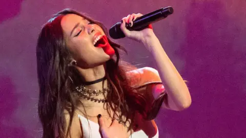 Getty image Olivia Rodrigo sings in a microphone, in which her hand is pressed on her chest and her eyes are closed