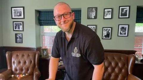 A man stands in a room with brown leather armchairs and smiles at the camera. He is bald and has a light-coloured beard.