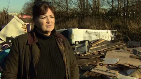 Kym Grant, a woman wearing a dark green jacket, stood by fly-tipped rubbish including fridges and bedframes