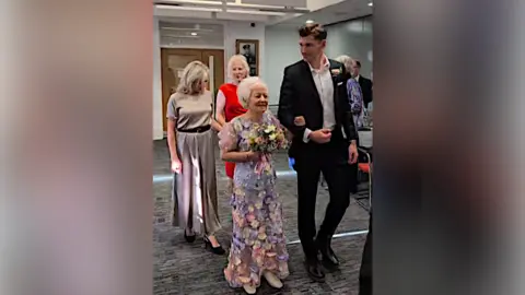 Contributed Jo is pictured walking down the aisle of a registry office with one arm looped in her grandson's. He wears a black suit with a white shirt underneath and he looks at his grandmother. Two women follow them behind. 