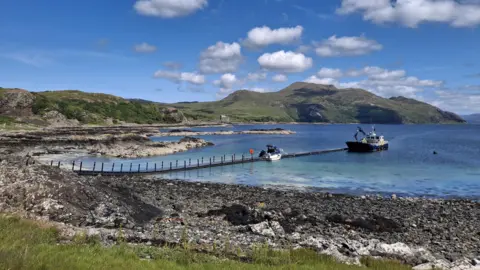 Ailie Surtees Clear blue water meets a stony beach. There is a small ferry at the end of a pier and a green hill in the background.