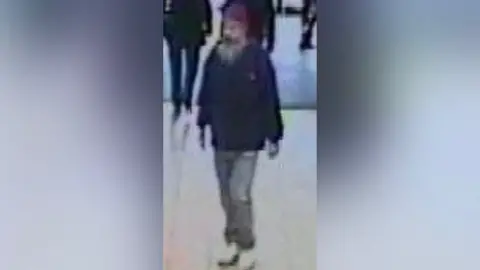 Locate International Man with grey beard wearing cap walks in a shopping centre with people behind him