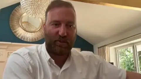 Scott El Paraiso, who is tanned and wearing a beard and a white shirt, sits looking into the camera on a Zoom call with leaseholders in June 2024