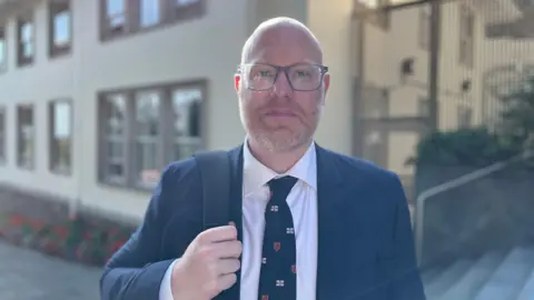 Deputy Rob Curgenven faces the camera. He is wearing a blue jacket, blue tie with flags of the Bailiwick of Guernsey on them and has a off white tie. He is wearing glasses and has a bag hanging from his right shoulder. 