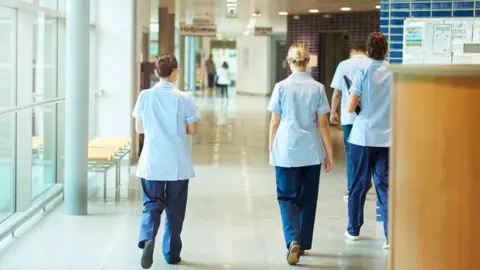 Getty Images Four nurses walk down a hospital corridor in a light blue uniform with dark blue trousers. The floor is white and they are beside a window.