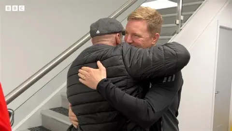 Steven hugging Eddie Howe. He is wearing a black coat and a grey cap. They are indoors by a flight of stairs