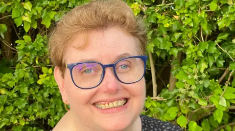 Amanda Cresswell A woman in her fifties, with short light brown hair and blue glasses smiles at camera.