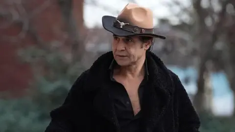 Chico arriving at court, wearing a brown cowboy hat with a silver cattle head adornment. He wears a black furry coat with a black open shirt underneath 