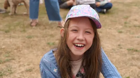 Stephen Huntley/BBC A young girl, smiling at the camera and wearing a hat with a blue jumper on. Other people are behind her, but you can only see their legs. Dried grass is behind her. 