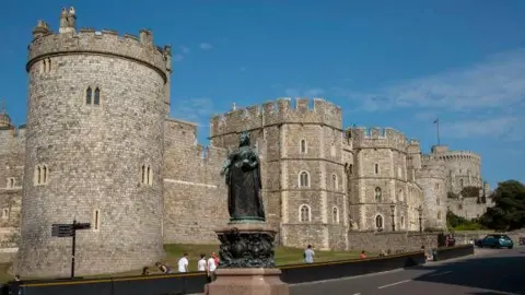 Getty Images Shot of the road outside the main entrance to Windsor Castle with the statue of Queen Victoria in the foreground and the Castle behind her with a bright sky above.