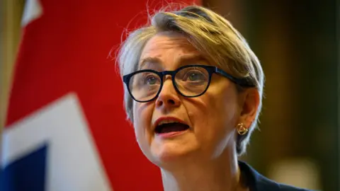 Foreign Secretary Yvette Cooper hosts a virtual meeting at the Foreign, Commonwealth and Development Office (FCDO) in Westminster. She wears black glasses, and a Union Jack is visible behind her.