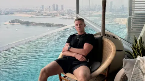 A man wearing a black shirt and shorts sits on a wicker chair next to a pool in a high-rise apartment. Behind him is a view of Dubai.