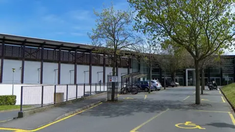 BBC Exterior of Beau Sejour leisure centre, a white and black building with trees at the front and several disabled car parking spaces