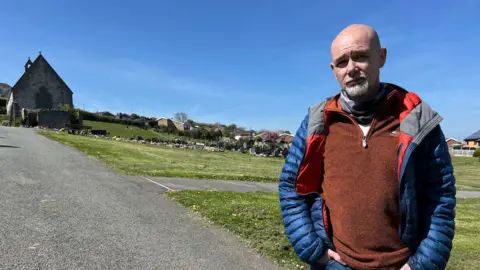A picture of David Pearson. He is standing at the forefront of Holywell cemetery, hands in jean pockets, looking at the camera. He wears an orange jumper and a blue jacket.
