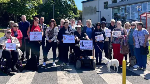 A group of men and women, the majority middle-aged or elderly, stand in the grounds of a leisure centre building in bright sunshine. One woman is sitting on a wheelchair, another in a mobility scooter. One holds a white dog on a lead. Some have placards with slogans reading: "Real trainers, not AI screens", and "Would you take 40% less income."