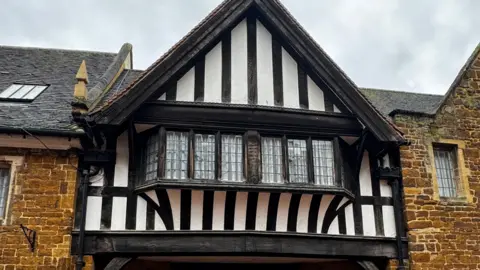 Prop-Search The outside of an old building, showing a wooden gable, with small windows on it and brick work around it. 