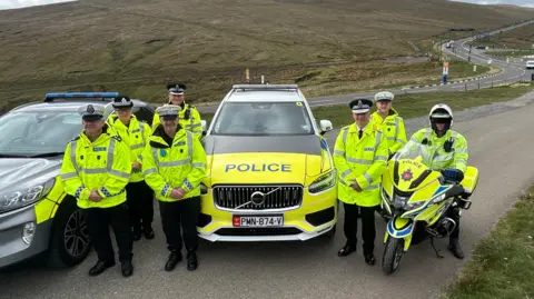 Seven police officers are positioned next to a police car, one is on a police bike. They are wearing hi-vis jackets and black trousers and boots and black and white hats. They stand next to the mountain road, part of the circuit TT riders race on during the festival.