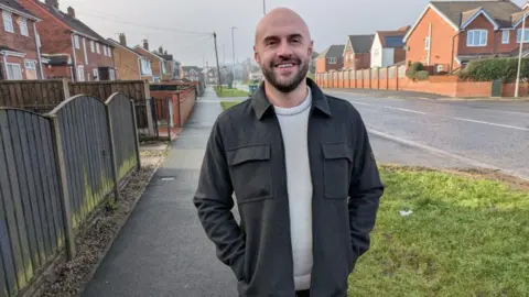 David Spereall/BBC A man in his thirties with a shaved head and a short beard. He is wearing a black jacket and cream sweater. He is stood on a pavement next to a suburban road and smiling at the camera.
