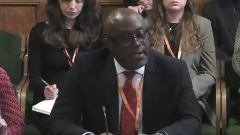 Parliamentlive.tv Long Lartin's governor sits in front of a committee of MPs in a room in the Houses of Parliament. He is wearing a black suit, red tie and orange lanyard