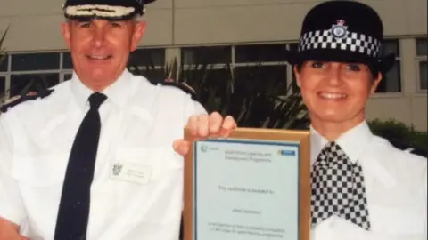 Janet Lovick Two police officers, one a man in a tie and and a braided hat, the other, a woman, wearing a chequered hat and neck tie, holding a certificate