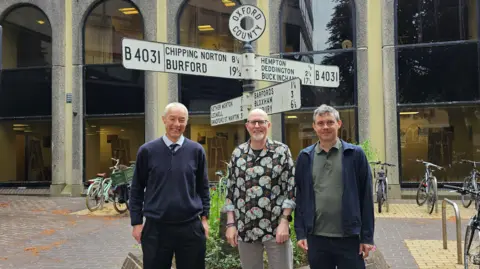 Pete Sudbury, Ian Middleton and Robin Bennett stand outside the county council offices. They are in front of location signs, but they are not in the modern style, and point in the directions of places like Burford, Ledwell, Buckingham, and Bloxham. Pete has short grey hair and wears a blue v-neck jumper over a tie and light blue shirt. Ian has a white goatee, wears black-rimmed glasses, and a black, grey and orange patterned shirt. Robin has short greying hair and wears a blue jacket over a green shirt.